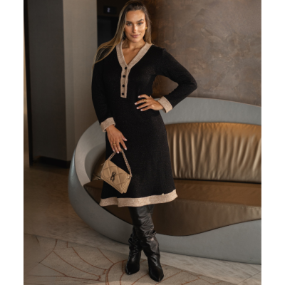 Mulher com vestido de malha preto com detalhes bege, botas pretas e mala bege num ambiente interior.