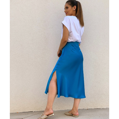 Mulher de costas a usar saia midi azul com corte lateral e t-shirt branca num fundo de parede clara