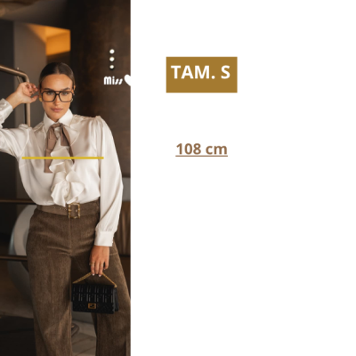 Mulher vestindo blusa branca com folhos e laço, calças castanhas e mala preta, texto TAM. S e 108 cm à direita