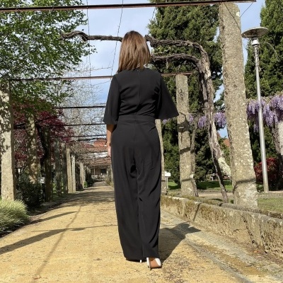 Mulher de costas com macacão preto em alameda com vegetação e colunas de pedra