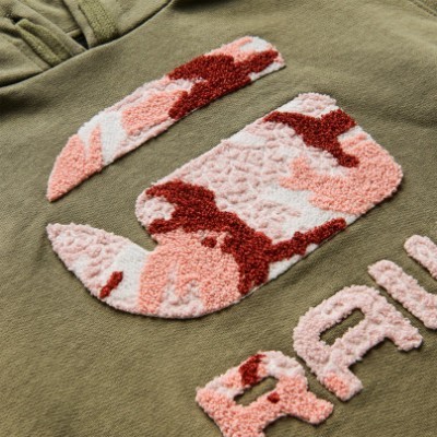 Detalhe de tecido verde-oliva com bordado colorido em rosa, vermelho e branco formando letras parciais.
