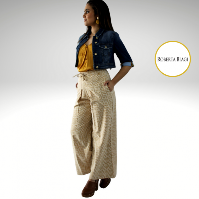 Mulher com casaco azul de ganga, blusa mostarda e calças largas bege com padrão geométrico, sapatos castanhos, fundo branco com logo ROBERTA BIAGI.