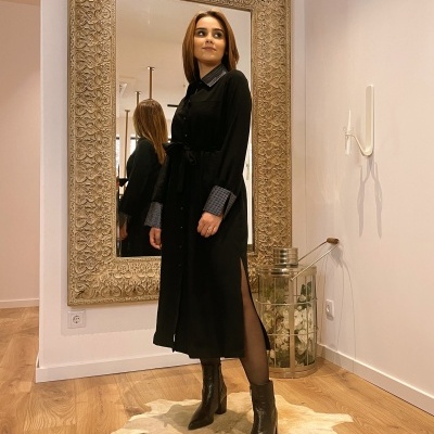 Mulher com vestido preto de padrão cinza nos punhos e gola, botas pretas, em interior com espelho grande e chão de madeira.