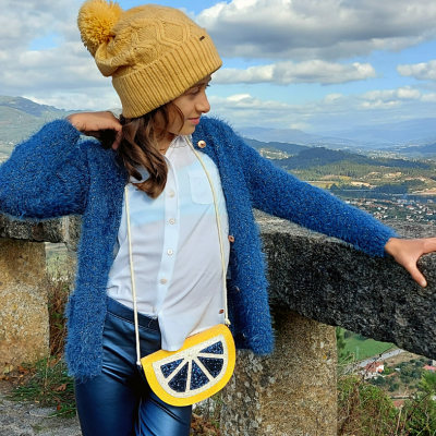 Mulher com casaco azul e gorro amarelo com bolsa em forma de limão num cenário de montanha.