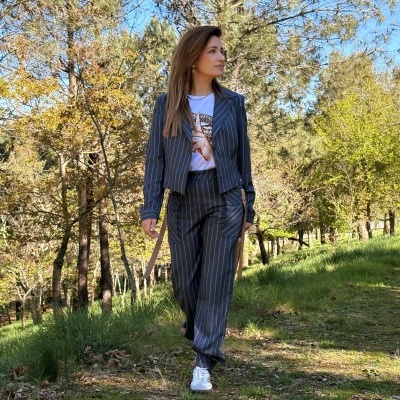 Mulher a usar conjunto risca de giz cinzento com blazer e calças, t-shirt branca e sapatilhas, numa zona exterior com árvores