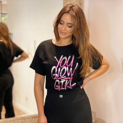 T-shirt preta com texto YOU GLOW GIRL em rosa e branco, usada por mulher num interior