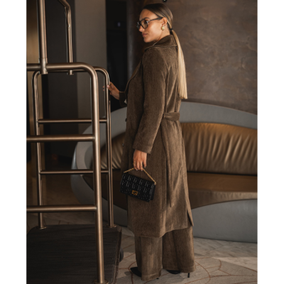 Mulher com casaco castanho longo e calças largas, segurando mala preta com corrente dourada, em ambiente interior com sofás e estrutura metálica.