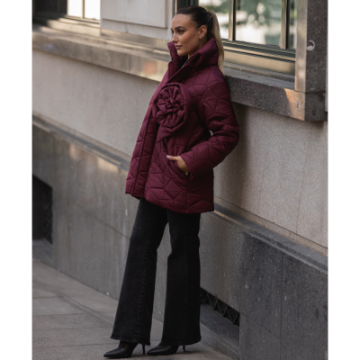 Mulher com casaco acolchoado bordeaux com flor na frente, calças pretas largas e sapatos de salto em ambiente urbano