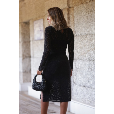 Mulher de vestido midi preto de renda segurando mala preta em ambiente com parede de pedra