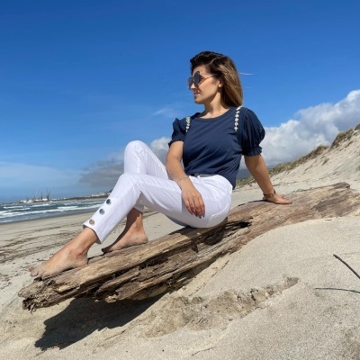 Mulher sentada numa praia de areia, com blusa azul-marinho e calças brancas, óculos de sol, céu azul e tronco de árvore.