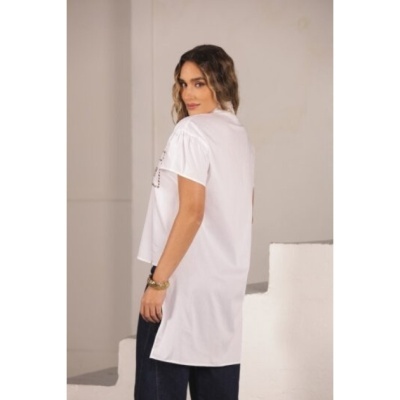 Mulher com t-shirt branca comprida com estampado preto e calças escuras, em ambiente interior claro.