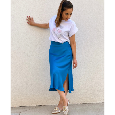 Mulher com t-shirt branca estampada de flores e saia azul com fenda encostada numa parede