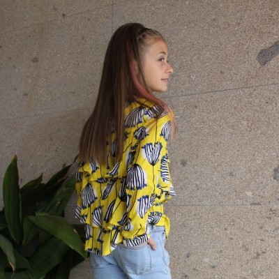 Menina com blusa amarela estampada e calças de ganga junto a parede de pedra