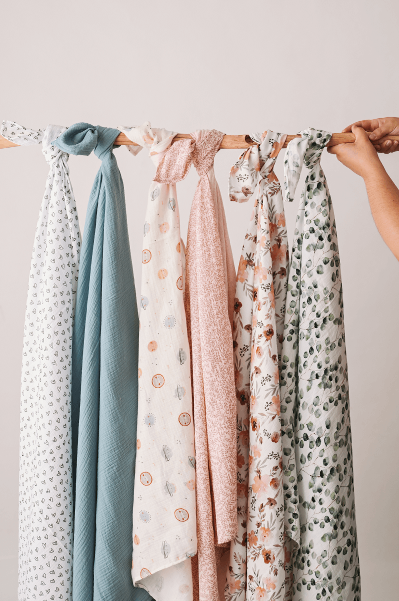 Fralda Swaddle Seis panos leves pendurados num varão de madeira com padrões florais e cores suaves.