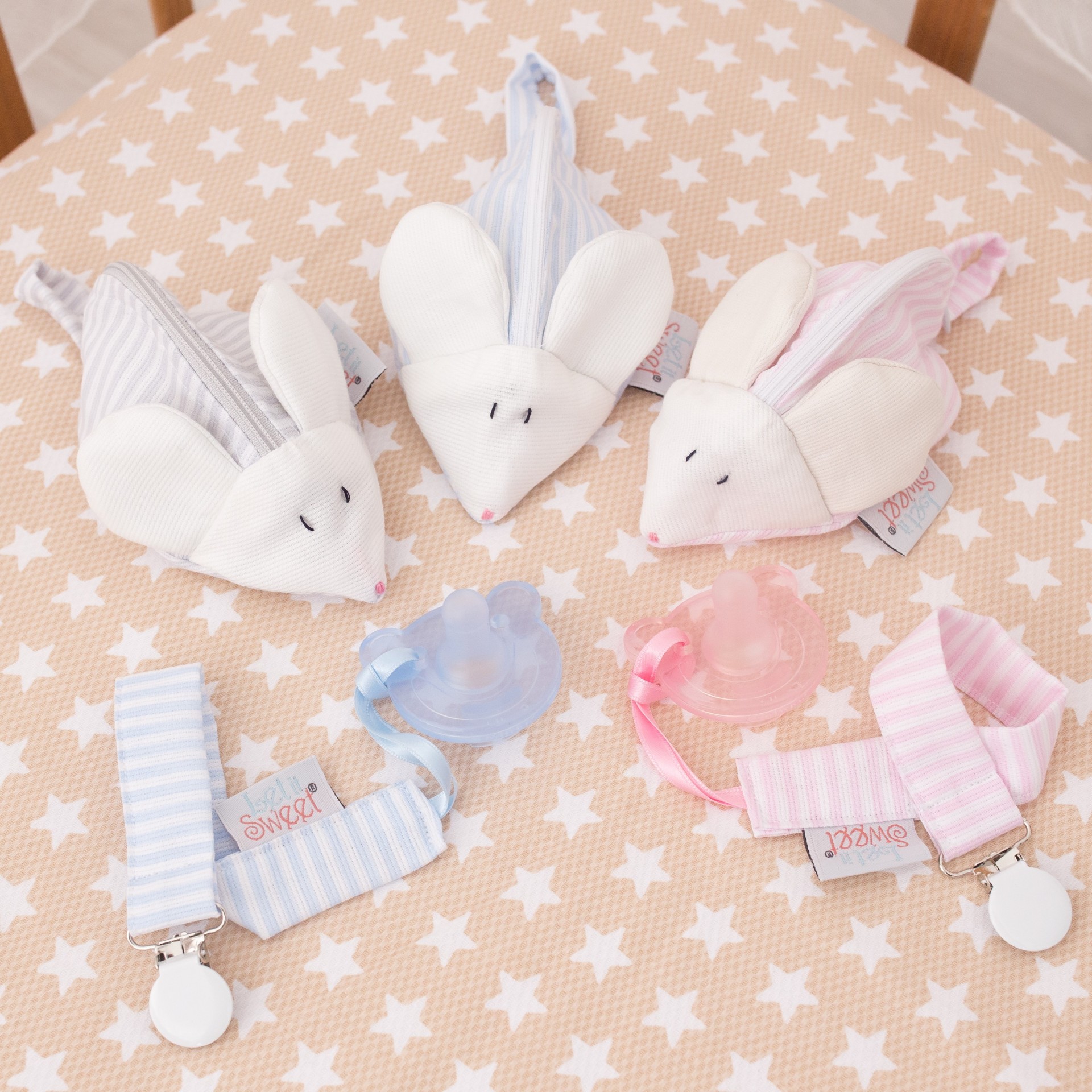 Fita para chupeta Chupetas transparentes azul, rosa e lilás com presilhas às riscas e brinquedos de pelúcia em forma de rato em fundo bege com estrelas brancas