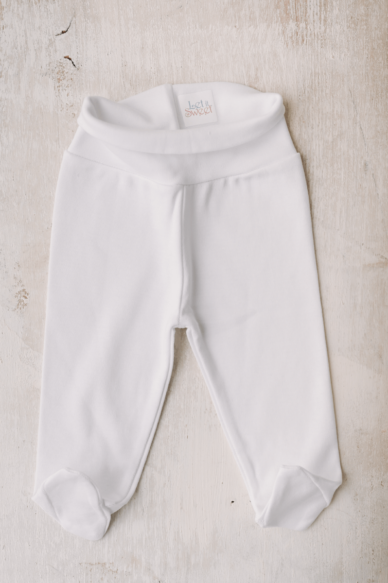 Conjunto de roupinha Calças brancas para bebé com pés fechados e etiqueta 'legit SWEET' em fundo de madeira clara
