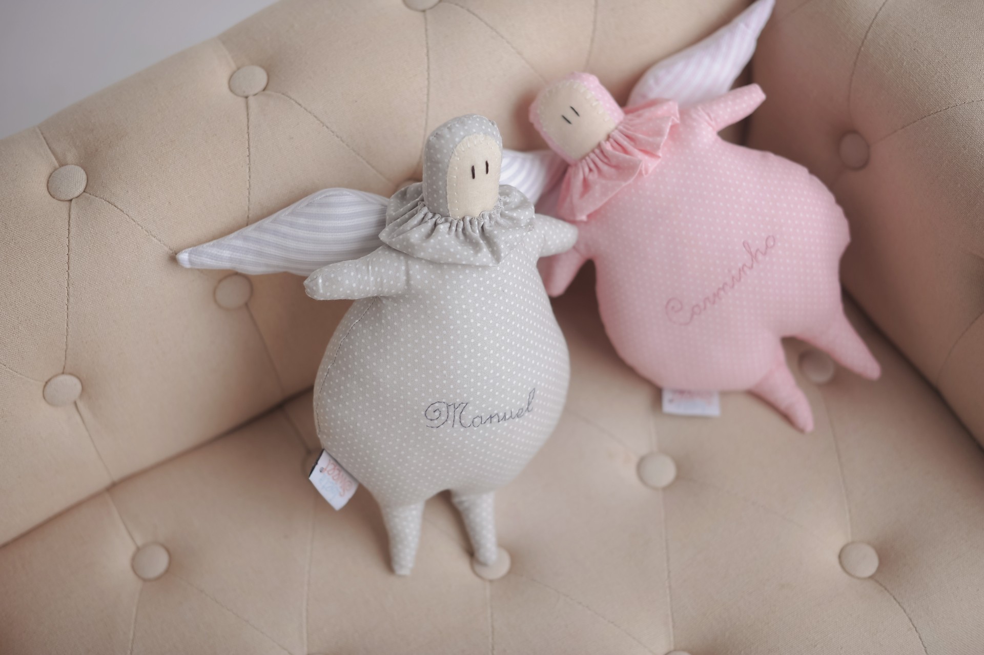 Anjinho Lemonade Dois bonecos de tecido em forma de anjos, cinzento e rosa, num sofá bege acolchoado
