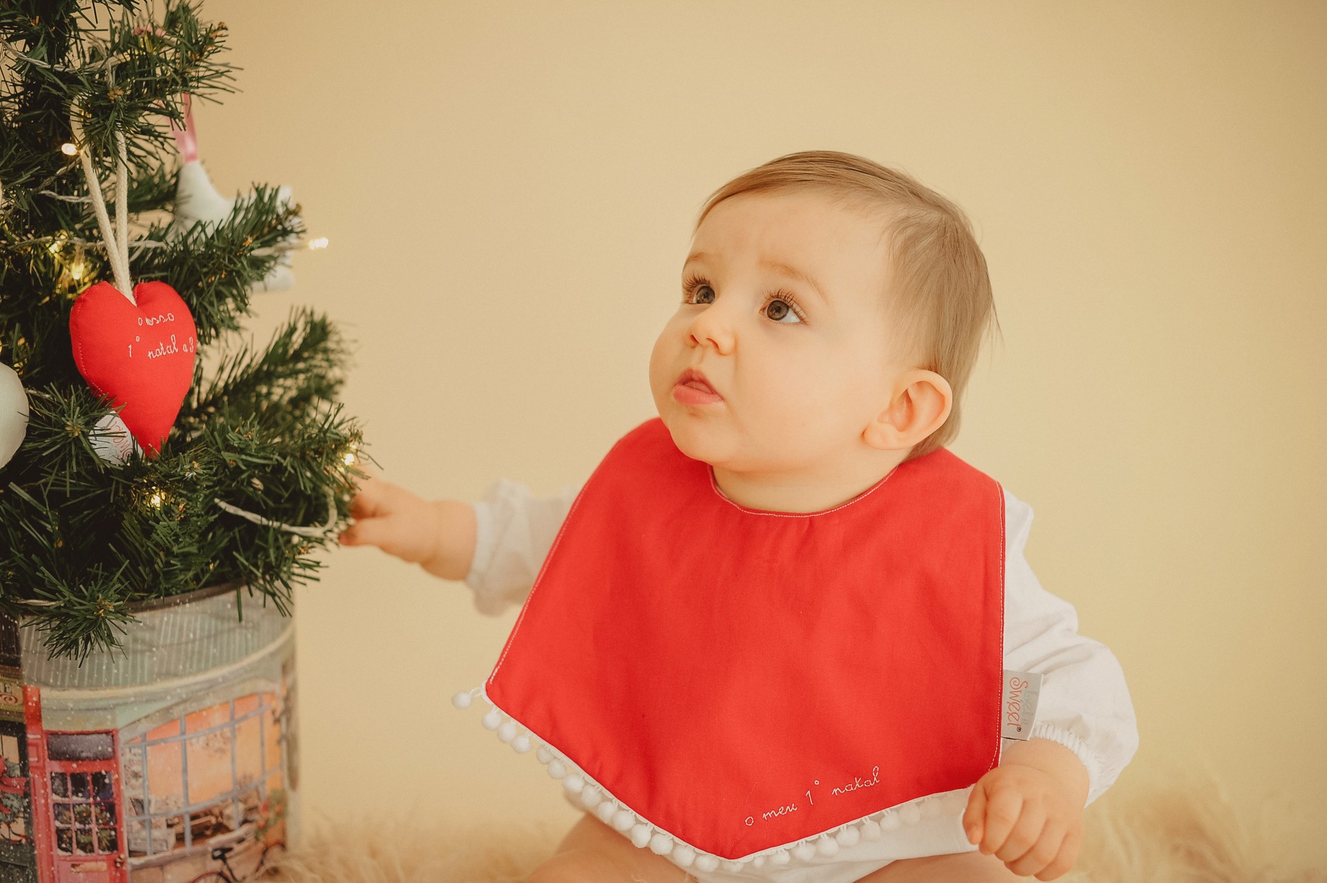 Babete de Natal Bebé com babete vermelho junto a árvore de Natal decorada