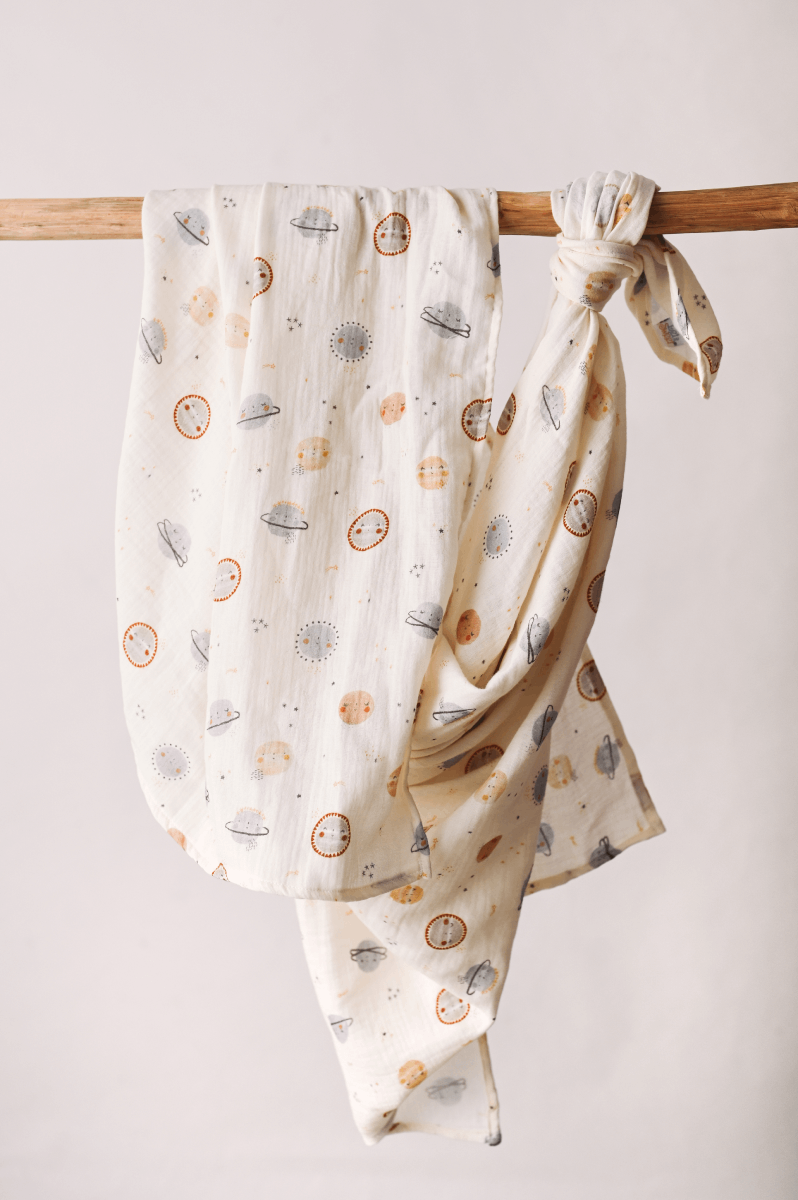 Fralda Swaddle tecido bege claro com estampados de planetas e sóis em várias cores pendurado num varão de madeira