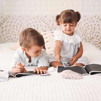 Duas crianças a desenhar em livros com páginas escuras numa cama branca com colcha matelassada.