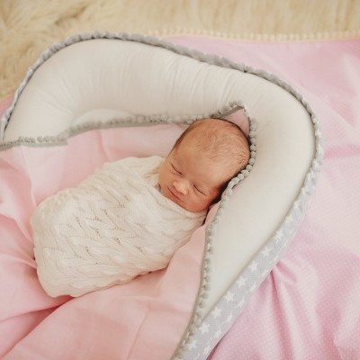 Bebé recém-nascido envolto em manta branca deitado em almofada cinza com estrelas, sobre cobertor rosa e tapete felpudo bege