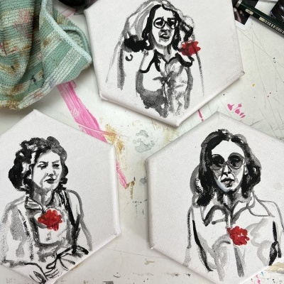 Três pinturas em telas hexagonais representando mulheres com flores vermelhas no peito sobre uma mesa branca com manchas de tinta