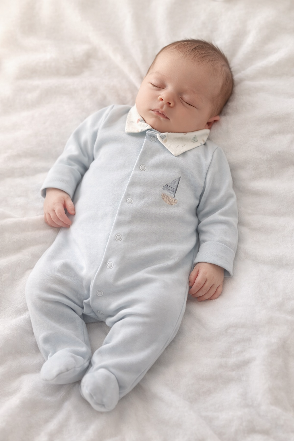Babygrow 'Blue Sea' com gola aberta Bebé a dormir com macacão cinzento claro de barco bordado e gola branca, deitado em manta branca