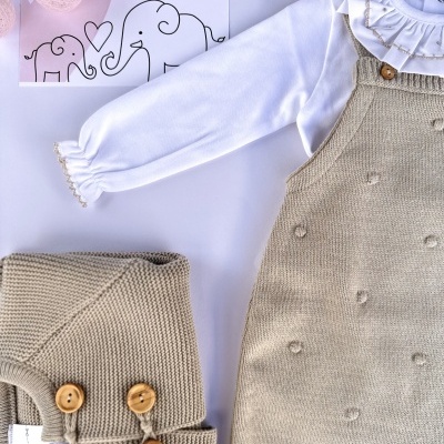 Conjunto de roupa de bebé com body branco e jardineira bege com botões de madeira, casaco bege de malha, sobre fundo claro com decoração de luzes