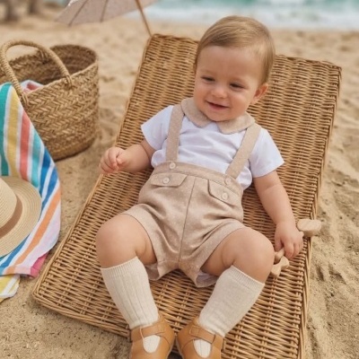 Bebé sentado numa espreguiçadeira na praia com roupas claras e sapatos castanhos.