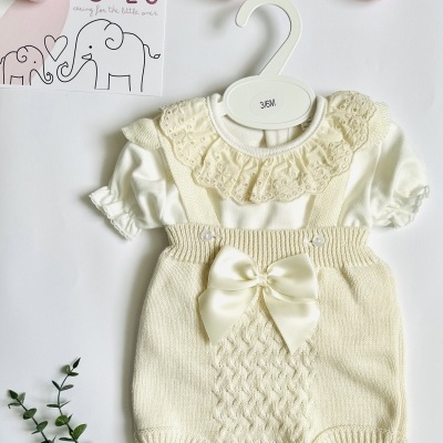 Roupa de bebé creme com blusa rendada e malha trançada sobre fundo branco