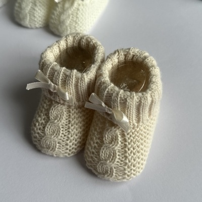 Sapatinhos de bebé de lã creme com padrão de trança e laço, sobre fundo branco