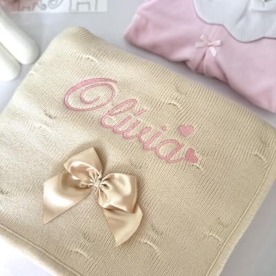 manta bege tricotada com nome Olívia bordado a rosa e laço de cetim, blusa rosa com gola branca ao fundo