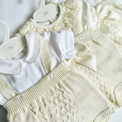 Conjunto de roupa de bebé creme e branco em malha com padrão trançado, exposto em cabides brancos
