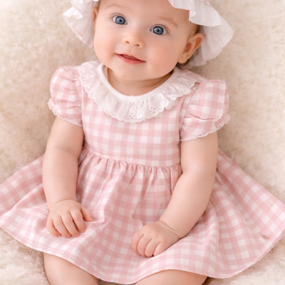 Bebé vestido com conjunto rosa e branco xadrez e chapéu, sentado sobre superfície macia branca