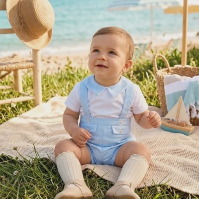 Bebé vestido de azul claro e branco sentado na relva junto à praia com acessórios de verão em redor.