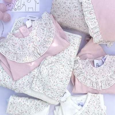 Conjunto de roupa de bebé rosa e branco com padrão floral pequeno e folhos, almofada e gorro, sobre fundo branco e luzinhas.