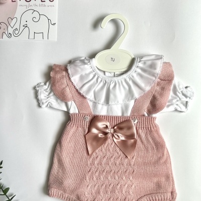 Conjunto de roupa de bebé branca e rosa com laço, pendurado em cabide branco.
