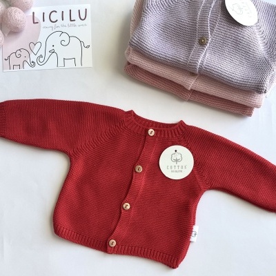Casaco de malha para bebé vermelho com botões de madeira e etiquetas, com casacos empilhados lilás e rosa ao lado e cartão ilustrado
