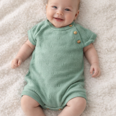 Bebé sorridente com roupinha verde de ponto e botões castanhos deitado em tecido felpudo branco