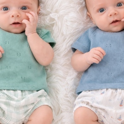 Dois bebés deitados lado a lado, um com camisa verde e cueiros xadrez, o outro com camisa azul e cueiros com desenhos