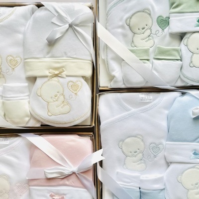 Conjuntos de roupa de bebé com ursinhos bordados e cores pastel, embalados em caixas com laços brancos.