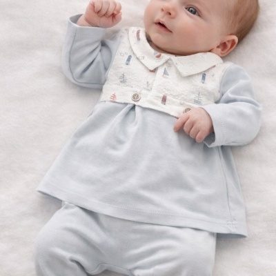 Bebé vestido com roupa azul clara e colete branco com padrão náutico a deitar-se numa superfície branca.