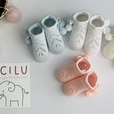 Sapatinhos de bebé em malha azul, branco, amarelo e rosa com pompons, junto a caixa com texto LICILU e luzes decorativas.