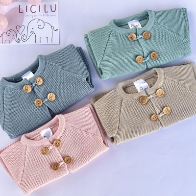 Quatro casacos de malha para bebé em cores pastel com botões de madeira sobre superfície branca e cartão decorativo com elefantes.