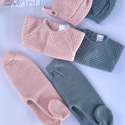 Conjunto de roupa de bebé rosa e cinza em malha texturizada com casacos, calças e gorros sobre fundo branco.