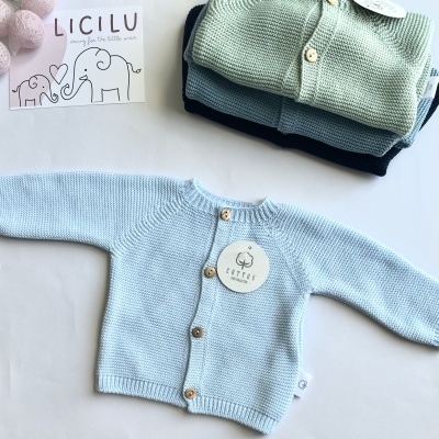 Casaco de malha azul claro para bebé com botões de madeira e etiqueta, junto a casacos dobrados e cartão LICILU