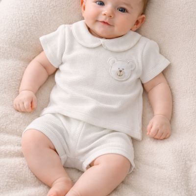 Bebé vestido com conjunto branco texturizado com ursinho bordado deitado sobre manta creme felpuda