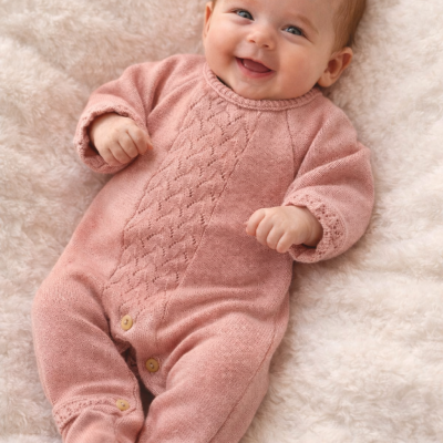 Bebé a sorrir de macacão rosa de malha com botões de madeira, deitado numa manta felpuda