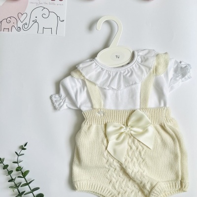 Conjunto de roupa de bebé com body branco e jardineiras creme com laço, apresentado em cabide branco sobre fundo branco