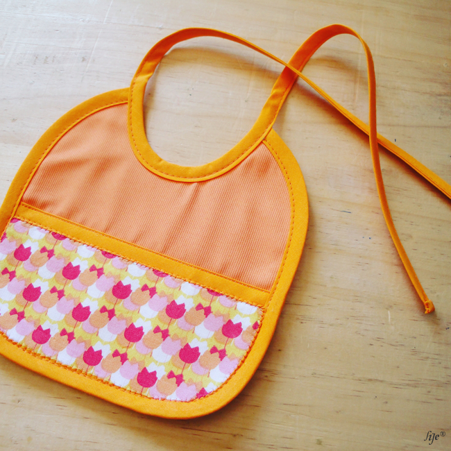 Babete para bebé laranja com padrão colorido sobre madeira clara